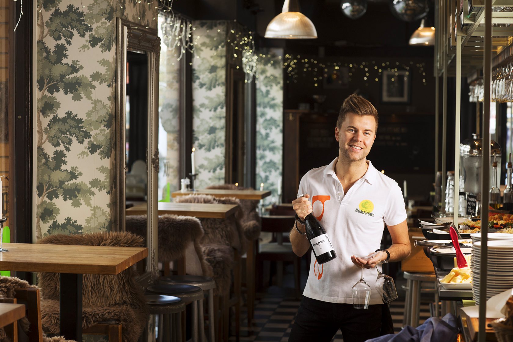 En man står i en restaurang och håller i en vinflaska och ett glas.