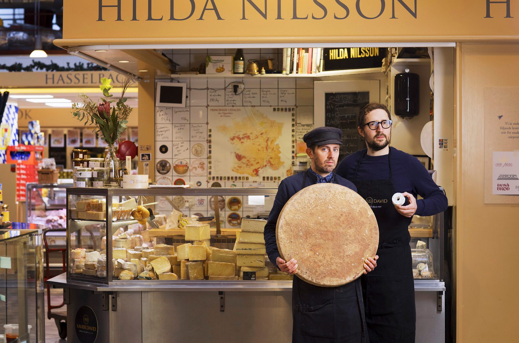 Två män står framför sin ostbutik med en stor och en liten ost i händerna.